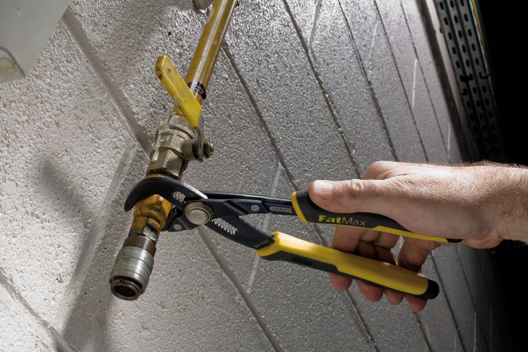 Una persona utiliza unas pinzas ajustables Stanley FatMax para sujetar y girar una unión de tubería conectada a una tubería amarilla junto a una pared blanca texturizada.