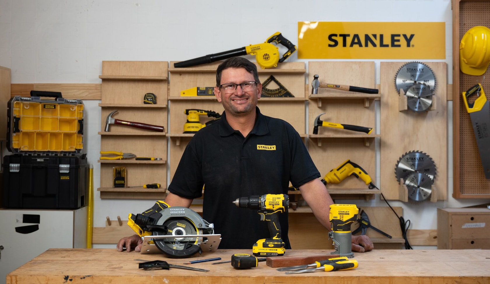 Una persona con una camiseta negra STANLEY está detrás de un banco de trabajo mostrando varias herramientas eléctricas y manuales STANLEY en amarillo y negro. El fondo del taller tiene estanterías con más herramientas STANLEY, cajas de herramientas, hojas de sierra y un casco de seguridad amarillo. La cara de la persona está desenfocada por privacidad.